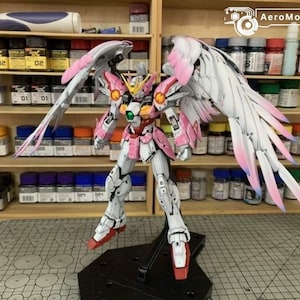 May include: A white and pink Wing Gundam model kit figure featuring large feathered wings with pink-tipped feathers, a yellow V-fin, and a green chest gem. The robot stands on a black display base, showcasing intricate panel lining and armor details.