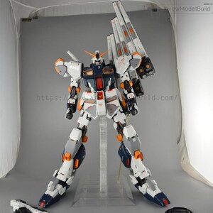 May include: A detailed model of a Gundam robot, primarily white, gray, and navy blue, with orange and red accents. The model is posed on a clear stand, showcasing its intricate design and mechanical details. The text "ArrowModelBuild" is visible.