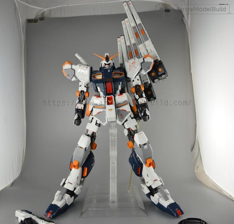 May include: A detailed model of a Gundam robot, primarily white, gray, and navy blue, with orange and red accents. The model is posed on a clear stand, showcasing its intricate design and mechanical details. The text "ArrowModelBuild" is visible.