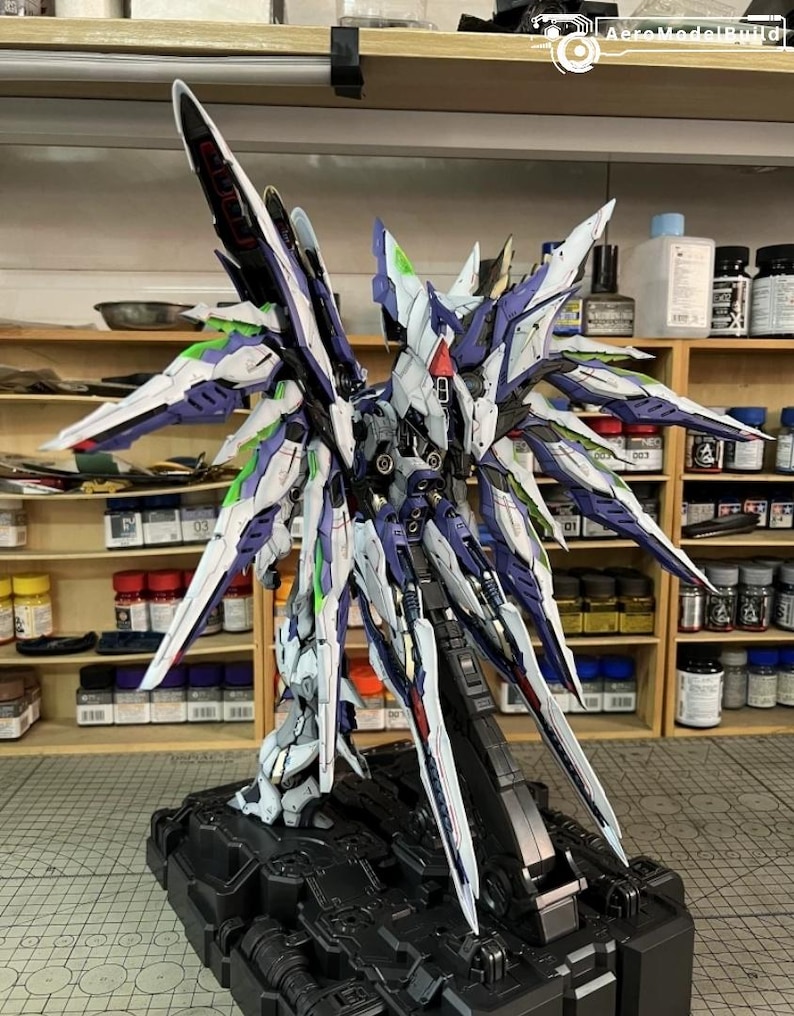 May include: A highly detailed model of a futuristic robot with large, stylized wings. The model features a complex design with white, blue, green, and red accents. It is mounted on a black base, showcasing intricate mechanical details.