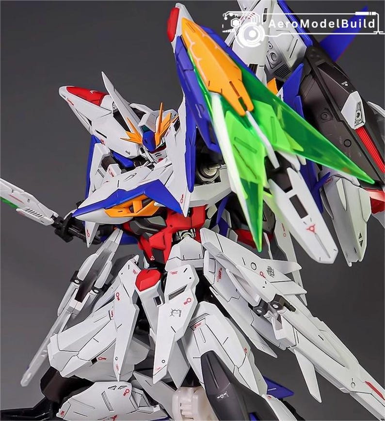 Puede incluir: Maqueta detallada de un robot futurista, principalmente blanco con detalles en azul, rojo y amarillo. El robot presenta paneles intrincados, bordes afilados y componentes transl&uacute;cidos verdes y naranjas. El logotipo de AeroModelBuild es visible.