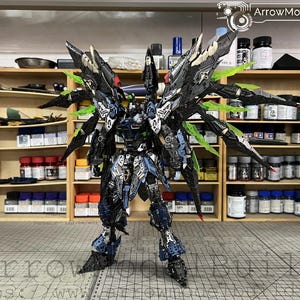 May include: A detailed model robot with black, blue, and green detailing. The robot has large wings and intricate features. The background shows shelves filled with paint bottles and supplies. The text "ArrowModelBuild" is visible.