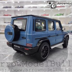 May include: A blue diecast model of a Mercedes-Benz G 63 AMG SUV. The model has black wheels, a spare tire on the back, and AMG badging. The background shows a display case with other model cars. The text "ArrowModelBuild" is at the bottom.
