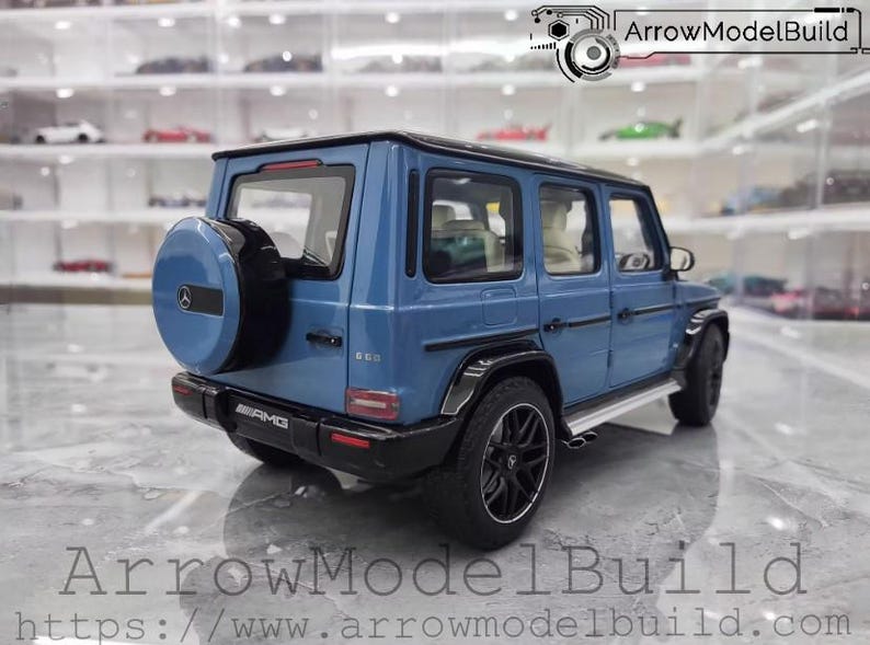 May include: A blue diecast model of a Mercedes-Benz G 63 AMG SUV. The model has black wheels, a spare tire on the back, and AMG badging. The background shows a display case with other model cars. The text "ArrowModelBuild" is at the bottom.