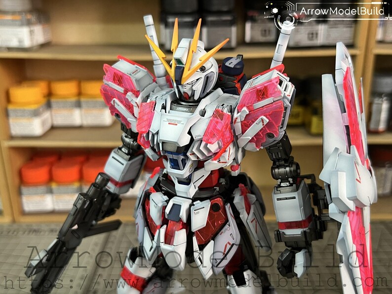 May include: A detailed model of a Gundam robot, primarily white, red, and pink. The robot has a complex design with multiple panels, weapons, and a shield. The model is posed in a dynamic stance, showcasing its intricate details.