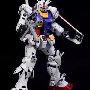 Op de afbeelding: Gedetailleerd model van een witte, blauwe, rode en gele Gundam-robot, geposeerd tegen een zwarte achtergrond. De robot heeft een geweer en een schild. Het model is een gedetailleerde weergave van een populair anime-personage.