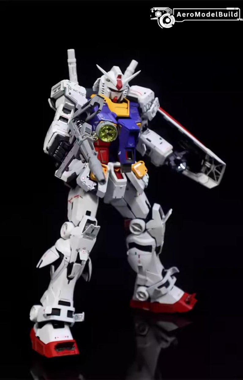 Op de afbeelding: Gedetailleerd model van een witte, blauwe, rode en gele Gundam-robot, geposeerd tegen een zwarte achtergrond. De robot heeft een geweer en een schild. Het model is een gedetailleerde weergave van een populair anime-personage.