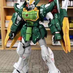 May include: A detailed model of a Gundam robot, predominantly green, white, and yellow. The robot has intricate armour plating, articulated joints, and a detailed head with yellow accents. The model is posed in a standing position, showcasing its design and construction.