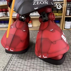 May include: Close-up of a red and black model robot's lower legs and feet. The model has detailed panel lines and white accents. The text "PRINCIPALITY OF ZEON" is visible on the black section. A yellow rod extends upwards.