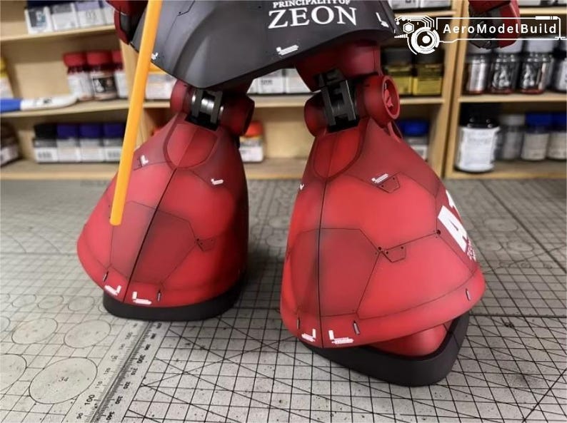 May include: Close-up of a red and black model robot's lower legs and feet. The model has detailed panel lines and white accents. The text "PRINCIPALITY OF ZEON" is visible on the black section. A yellow rod extends upwards.