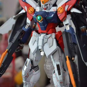 May include: A detailed model of a Gundam robot, featuring a color scheme of white, red, blue, and orange. The robot has large wings, two black guns, and intricate mechanical details. The model is standing on a clear plastic stand.