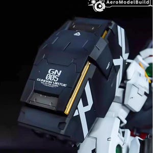 May include: A close-up of a black and white Gundam model kit. The model features a large, angular shield with gold accents and the text "GN 005 GUNDAM VIRTUE CELESTIAL BEING". The model is partially assembled.