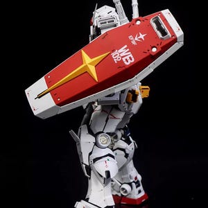 Op de afbeelding: Gedetailleerd model van een witte en rode robot met een groot schild. Het schild is rood met een gele ster en witte accenten. De robot heeft ingewikkelde mechanische details en de tekst "WB 102" is zichtbaar.