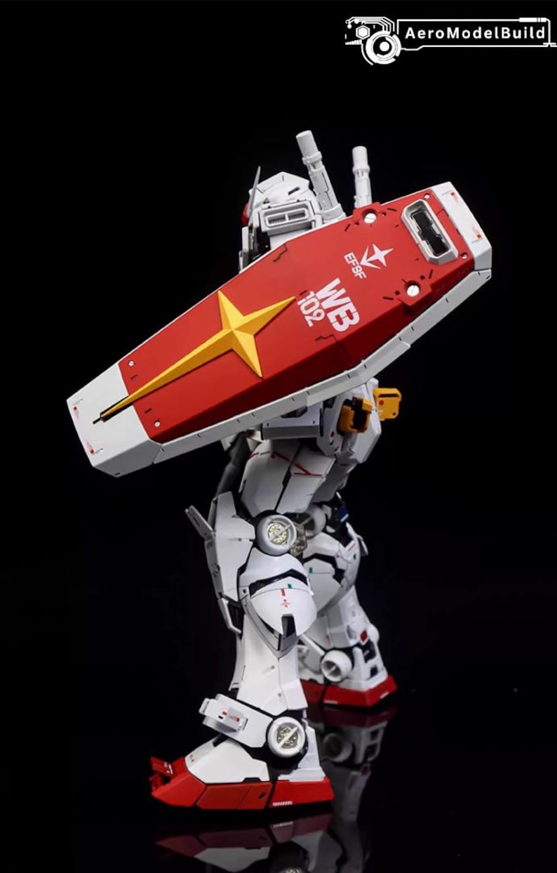 Op de afbeelding: Gedetailleerd model van een witte en rode robot met een groot schild. Het schild is rood met een gele ster en witte accenten. De robot heeft ingewikkelde mechanische details en de tekst "WB 102" is zichtbaar.