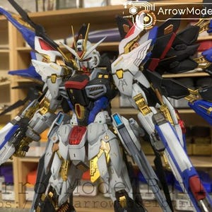 Puede incluir: Modelo detallado de un robot Gundam, con una combinaci&oacute;n de colores blanco, azul, rojo, negro y dorado. El modelo presenta intrincados detalles mec&aacute;nicos, alas y armas. El texto "ArrowModelBuild" es visible.