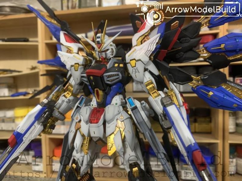 Puede incluir: Modelo detallado de un robot Gundam, con una combinaci&oacute;n de colores blanco, azul, rojo, negro y dorado. El modelo presenta intrincados detalles mec&aacute;nicos, alas y armas. El texto "ArrowModelBuild" es visible.