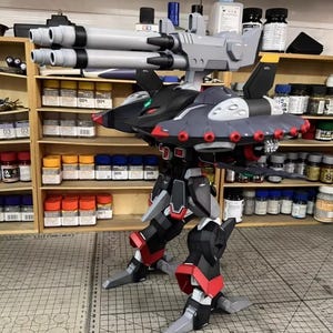 May include: AeroModelBuild model kit of a futuristic robot with a gray, black, and red color scheme. The robot has large cannons, wings, and detailed mechanical features. The model is assembled and stands on a cutting mat.