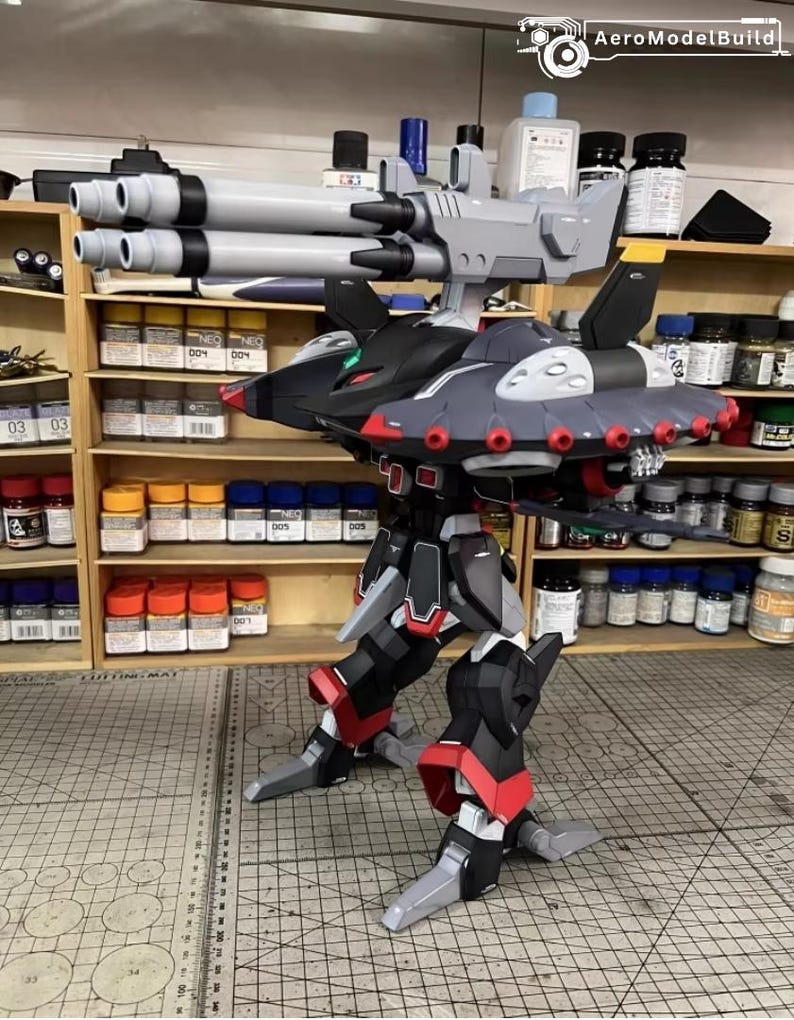 May include: AeroModelBuild model kit of a futuristic robot with a gray, black, and red color scheme. The robot has large cannons, wings, and detailed mechanical features. The model is assembled and stands on a cutting mat.