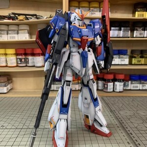 May include: A detailed model of a Gundam robot, predominantly white, blue, and red. The robot is posed with a large rifle and a red shield. The model features intricate panel lines and markings, showcasing a high level of detail.