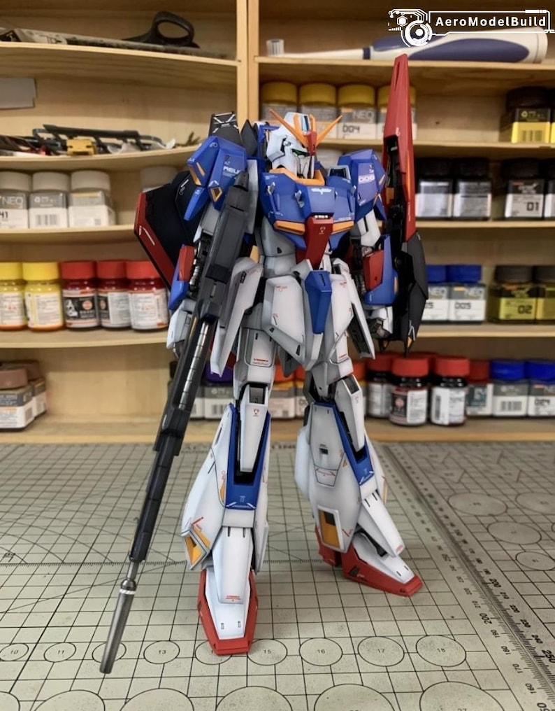 May include: A detailed model of a Gundam robot, predominantly white, blue, and red. The robot is posed with a large rifle and a red shield. The model features intricate panel lines and markings, showcasing a high level of detail.