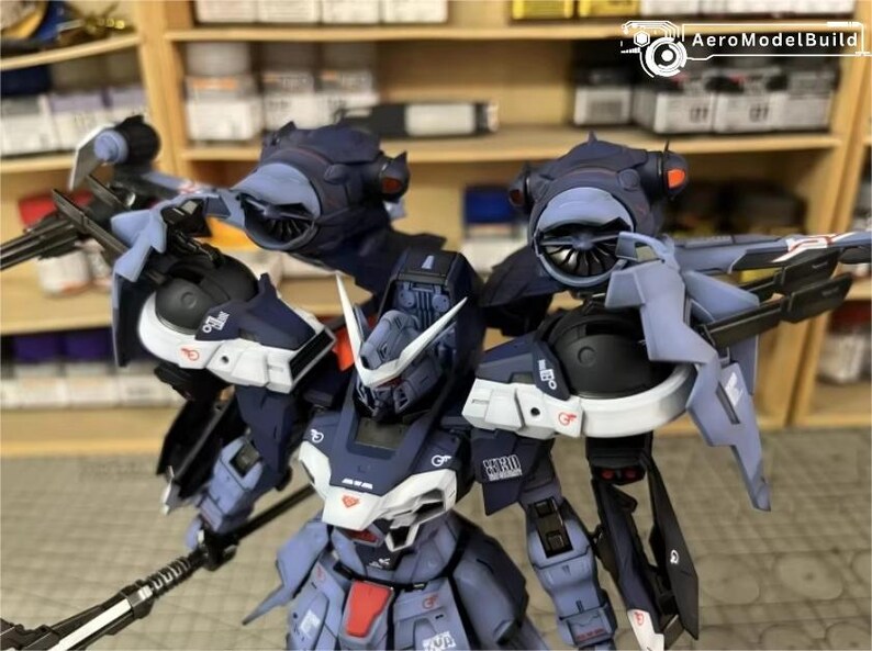 May include: A detailed model of a futuristic robot, primarily in shades of blue, gray, and white. The robot has intricate mechanical details, including wings, turbines, and a weapon. The model is set against a backdrop of shelves filled with various items.