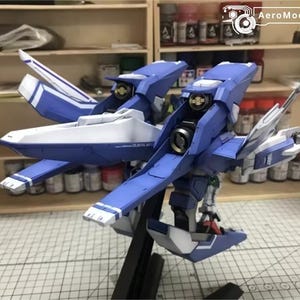 May include: A detailed model of a blue and white aircraft, featuring intricate wing and body designs. The model is mounted on a black stand, showcasing its complex structure. The background includes shelves with paint bottles and tools.