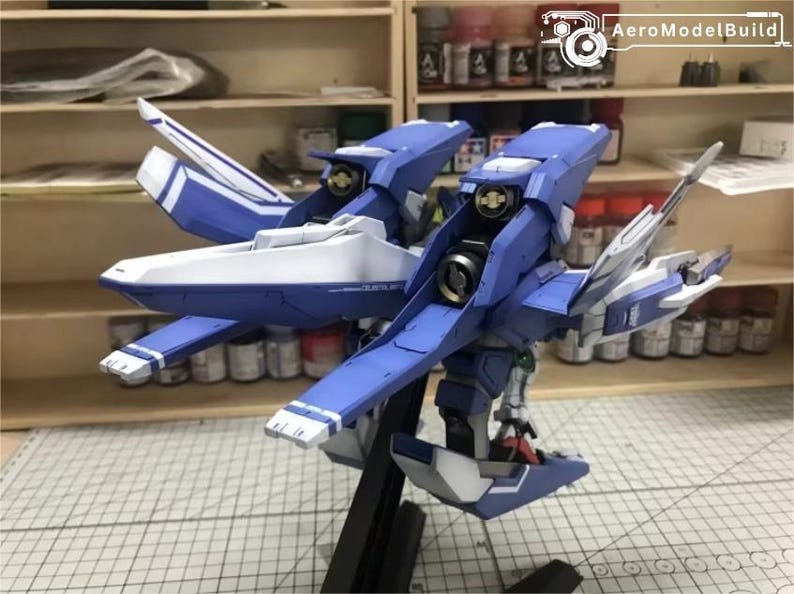 May include: A detailed model of a blue and white aircraft, featuring intricate wing and body designs. The model is mounted on a black stand, showcasing its complex structure. The background includes shelves with paint bottles and tools.
