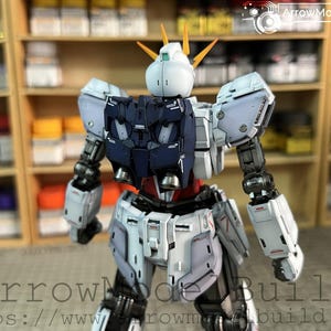 May include: A detailed model of a Gundam robot, primarily white, navy, and red, with intricate mechanical details. The model has a distinctive head with yellow accents and is posed in a dynamic stance. The background includes shelves with paint supplies.