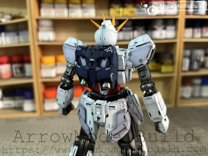 May include: A detailed model of a Gundam robot, primarily white, navy, and red, with intricate mechanical details. The model has a distinctive head with yellow accents and is posed in a dynamic stance. The background includes shelves with paint supplies.