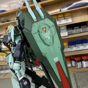 Puede incluir: Modelo detallado de un robot mecha, principalmente en tonos verdes, con detalles en rojo y negro. El robot presenta un gran escudo con una franja roja y detalles intrincados. El modelo se encuentra sobre un fondo de taller.