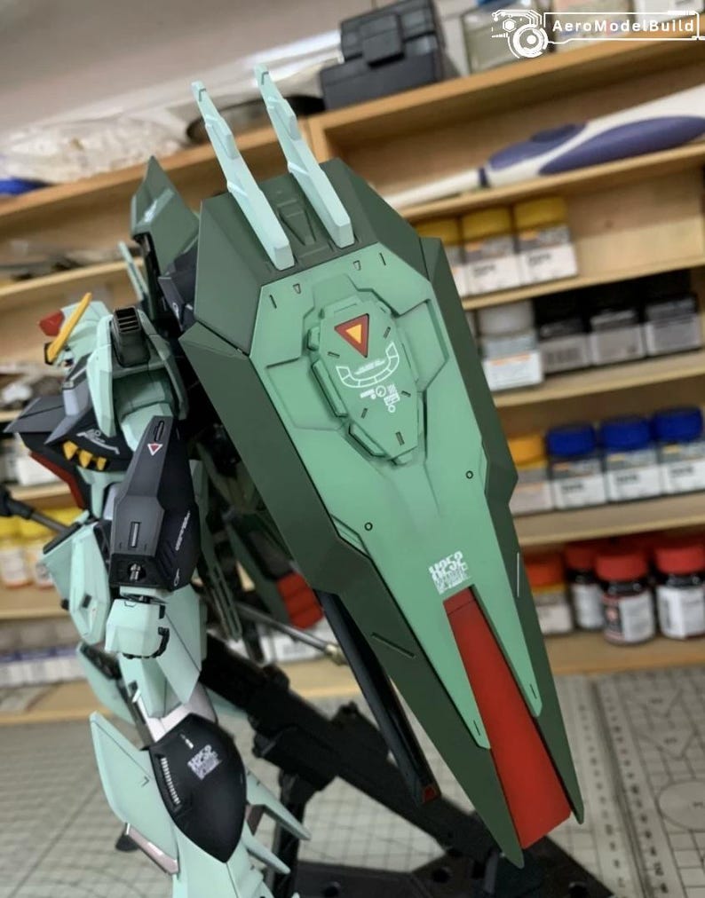Puede incluir: Modelo detallado de un robot mecha, principalmente en tonos verdes, con detalles en rojo y negro. El robot presenta un gran escudo con una franja roja y detalles intrincados. El modelo se encuentra sobre un fondo de taller.