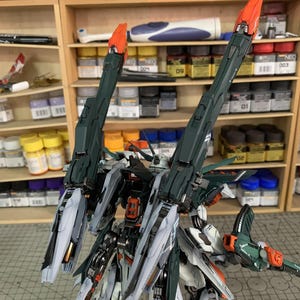 May include: A detailed model kit of a mech robot, featuring a complex design with green, white, and orange accents. The robot has large, angular wings and intricate mechanical details. The background includes shelves with paint supplies and tools.