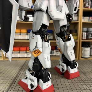 May include: A detailed model of a white and gray robot with red accents on the feet. The robot has intricate mechanical details, including visible joints and panels. The model is partially assembled, showcasing its internal structure. The text "AEUG" is visible on the robot's leg.