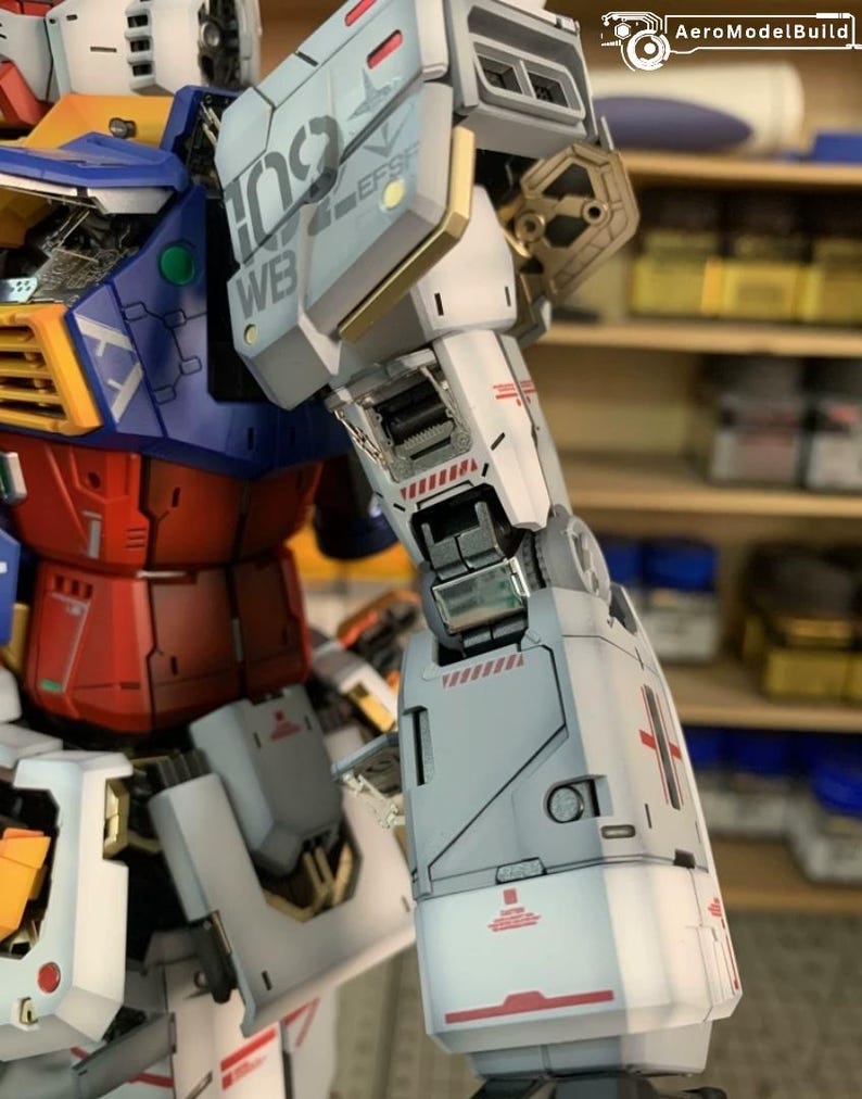 K&ouml;nnte beinhalten: Nahaufnahme eines detaillierten Modellroboters mit wei&szlig;em, rotem, blauem und goldenem Farbschema. Der Roboterarm weist komplizierte mechanische Details auf, einschlie&szlig;lich Tafeln mit Text und roten Akzenten. Das Modell ist teilweise montiert.