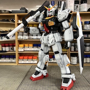 May include: A detailed model of a Gundam robot, primarily white with black, red, and yellow accents. The robot is holding a large weapon and has a shield. The model is standing on a gray surface with a grid pattern. The text "ArrowModelBuild" is visible.