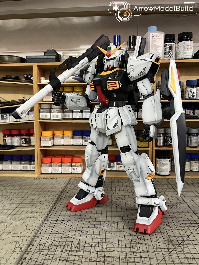 May include: A detailed model of a Gundam robot, primarily white with black, red, and yellow accents. The robot is holding a large weapon and has a shield. The model is standing on a gray surface with a grid pattern. The text "ArrowModelBuild" is visible.