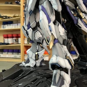May include: A close-up of a model robot, showcasing its complex mechanical design. The robot is predominantly white, grey, gold, and purple. It is positioned on a black, textured platform. The lighting accentuates the model's detailed construction.