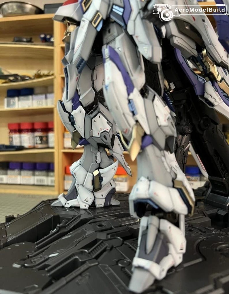 May include: A close-up of a model robot, showcasing its complex mechanical design. The robot is predominantly white, grey, gold, and purple. It is positioned on a black, textured platform. The lighting accentuates the model's detailed construction.
