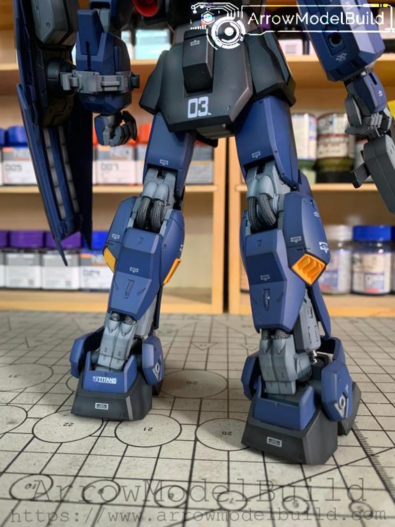 May include: A detailed, partially assembled model kit of a robot, primarily in shades of blue and gray. The model features intricate mechanical details, including segmented legs, with the number "03" visible. The word "TITANS" is printed on the foot.