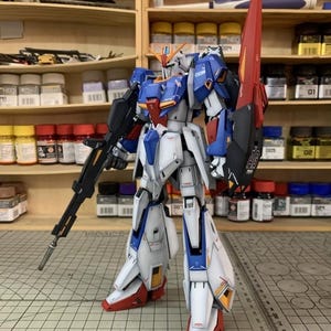 May include: A detailed model robot, predominantly white, blue, and red, holding a large rifle and shield. The robot is positioned on a grid surface, with shelves of paint and supplies in the background. The model is highly detailed.