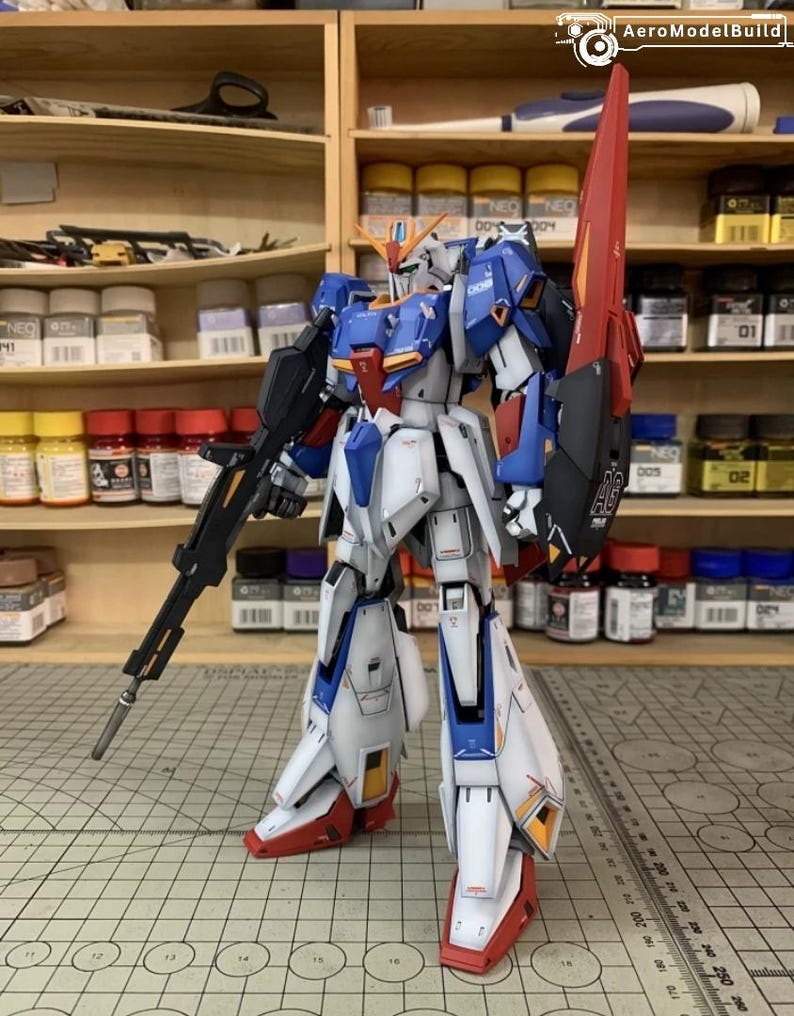May include: A detailed model robot, predominantly white, blue, and red, holding a large rifle and shield. The robot is positioned on a grid surface, with shelves of paint and supplies in the background. The model is highly detailed.