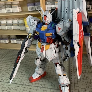 K&ouml;nnte beinhalten: Detailliertes Modell eines Gundam-Roboters mit einer Farbgebung aus Wei&szlig;, Blau, Rot und Gelb. Das Modell ist mit einem gro&szlig;en Schild, einem Gewehr und mehreren beweglichen Teilen ausgestattet. Das Modell steht auf einer Schneidematte.