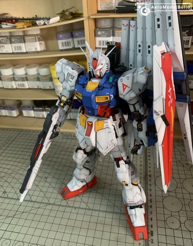 K&ouml;nnte beinhalten: Detailliertes Modell eines Gundam-Roboters mit einer Farbgebung aus Wei&szlig;, Blau, Rot und Gelb. Das Modell ist mit einem gro&szlig;en Schild, einem Gewehr und mehreren beweglichen Teilen ausgestattet. Das Modell steht auf einer Schneidematte.