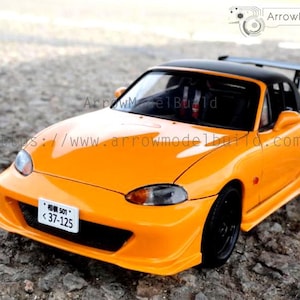Könnte beinhalten: Ein orangefarbenes Modellauto mit schwarzem Dach und Spoiler. Das Auto hat schwarze Räder und ein Nummernschild mit japanischen Schriftzeichen und Zahlen. Das Auto steht auf einer strukturierten Oberfläche.