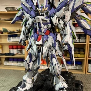 May include: A highly detailed model robot, featuring a colour scheme of white, blue, and grey, with elaborate wing-like components. The robot is positioned on a black base, highlighting its complex mechanical design. The image includes the text "AeroModelBuild".