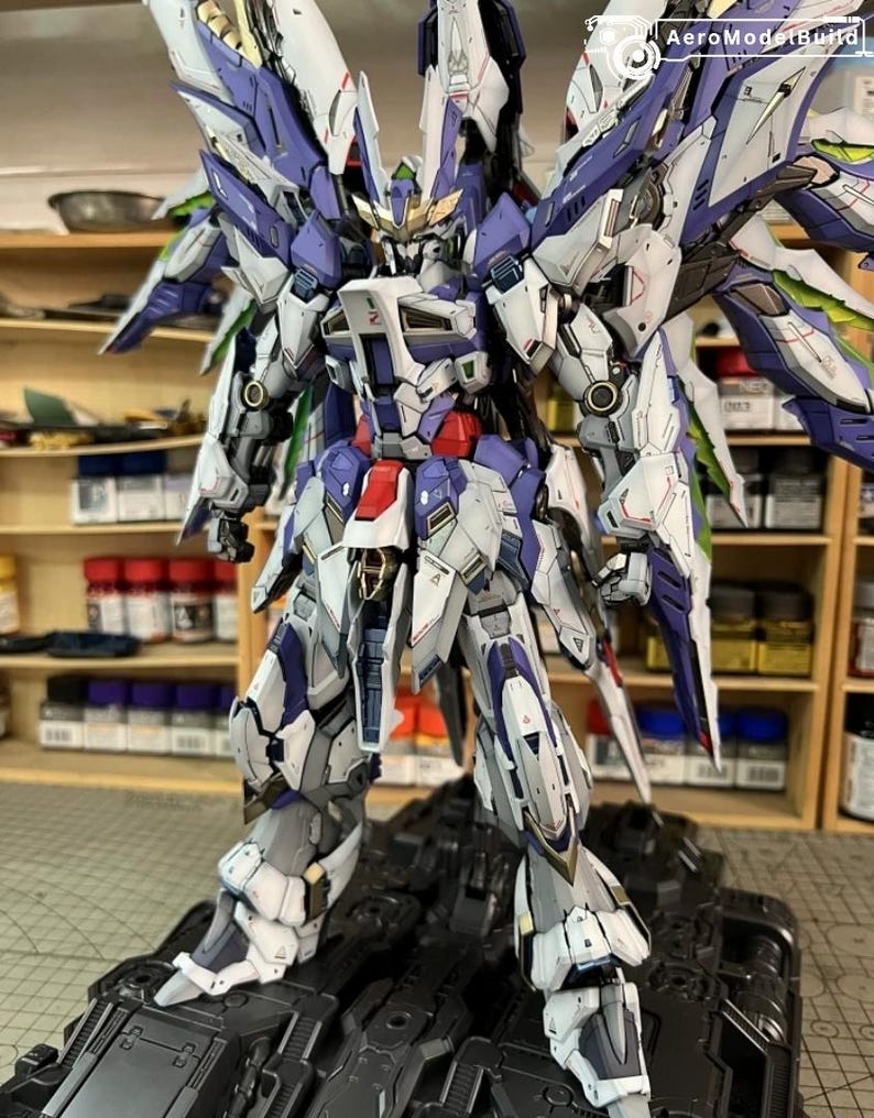 May include: A highly detailed model robot, featuring a colour scheme of white, blue, and grey, with elaborate wing-like components. The robot is positioned on a black base, highlighting its complex mechanical design. The image includes the text "AeroModelBuild".