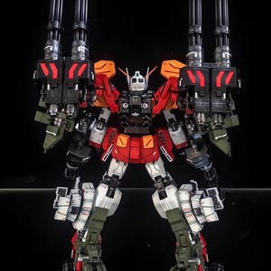 May include: A detailed model robot in red, white, black, and green. The robot has multiple cannons and weapons attached to its shoulders and back. The AeroModelBuild logo is in the top right corner.