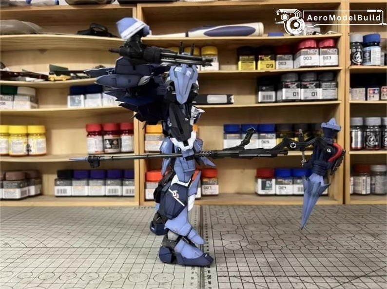 May include: A detailed model of a futuristic robot, primarily in shades of blue and black, with intricate mechanical details. The robot is posed with a long weapon, and the background shows shelves with paint bottles and model-building supplies.