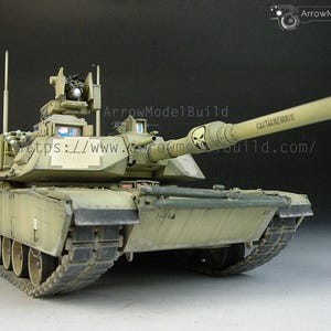 May include: A detailed model of a military tank in a desert camouflage colour scheme. The tank features a long barrel with the word "CONTAINEROUS" and a skull graphic. The model includes intricate details of the turret, tracks, and other components.
