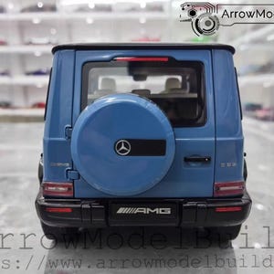 May include: A blue model car of a Mercedes-Benz G 63 AMG, viewed from the rear. The spare tire cover features the Mercedes-Benz logo. The car has black accents and the AMG logo is visible. The text "ArrowModelBuild" and a website address are at the bottom.